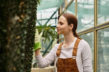 Genç bir kadın, yemyeşil çimlerle çevrili, sıcak bir serada bitki yetiştirmekten zevk alır..
