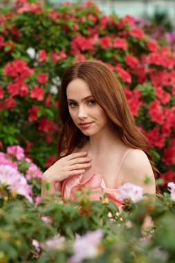Genç bir kadın renkli çiçeklerle çevrili yemyeşil bir serada duruyor zarafet ve cazibe saçıyor..