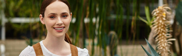 В ярко освещенной оранжерее молодая женщина улыбается в окружении пышных растений и листвы.