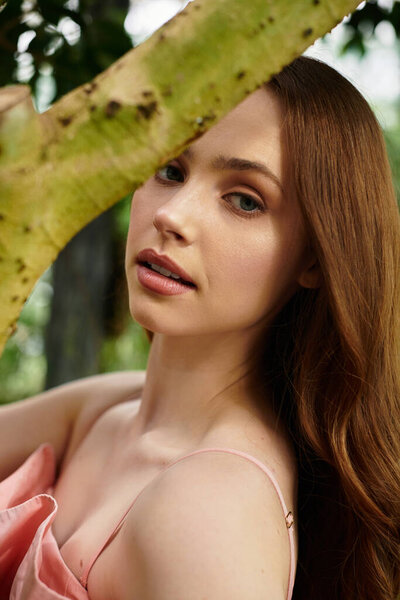 Молодая женщина с длинными волосами тихо улыбается, окруженная тропическими растениями в оранжерее.