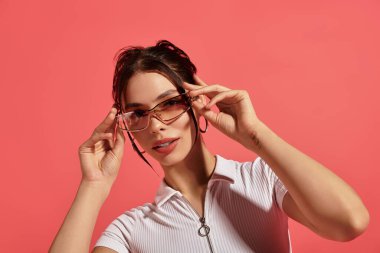 Çekici genç kadın, şık güneş gözlüklerini parlak bir ortamda ayarlarken kendine güvenerek poz veriyor..