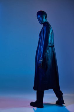 Genç bir Afro-Amerikan erkeği kendine güvenen şık bir şekilde duruyor, parlak mavi ve mor ışıklarla aydınlatılıyor..