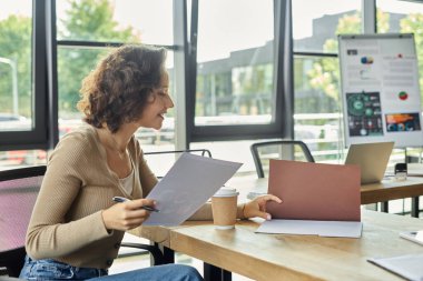 Bir kadın, modern ofis unsurları ve yeşilliklerle çevrili masasında düşünceli bir işe girişir..