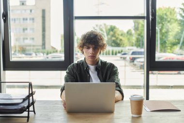 Genç bir insan kahve içerken dizüstü bilgisayarına odaklanmış bir şekilde masada oturuyor..