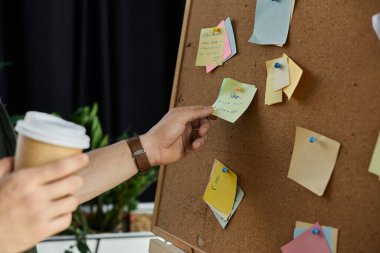 Bir kişi elinde kahve fincanı tutarken mantar panosundaki renkli yapışkan notları inceliyor..
