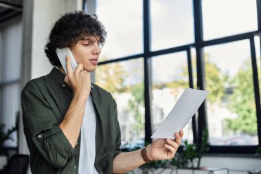 Telefondaki kişi, elinde kağıtlar, modern ofis ortamında kendini adamış biri..