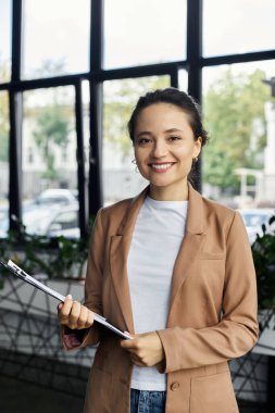 Modern bir ofiste genç bir profesyonel elinde bir dosya tutarken kendinden emin bir şekilde gülümsüyor..