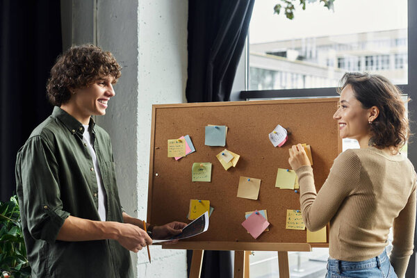 Двое коллег с радостью делятся идеями вокруг пробковой доски, заполненной красочными нотами.