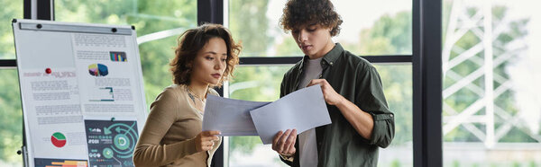 Two colleagues discuss project details while reviewing documents in a bright office environment.