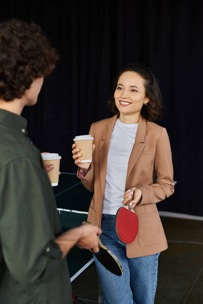 Two colleagues enjoy a lively chat while holding coffee cups and ping pong paddles in an office.