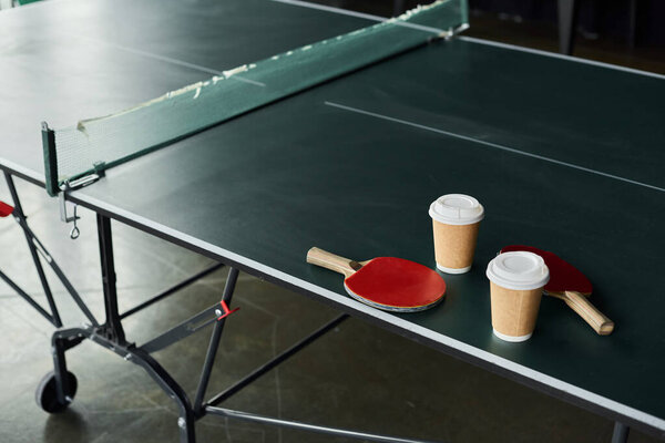Two paddles and coffee cups rest on a table next to a ping pong table during a work break.