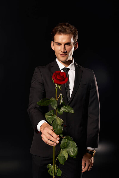 A young man stands confidently in a sleek black suit, offering a vibrant red rose with charm.