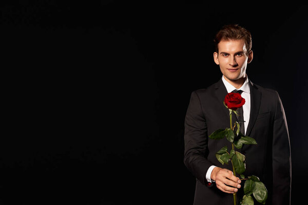 A dapper man stands confidently in formal attire, offering a stunning red rose with charm.