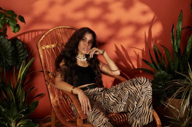 Genç bir kadın yeşil ve sıcacık renklerle çevrili bir sandalyede zarifçe oturuyor..