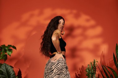 Göz kamaştırıcı genç bir kadın, canlı yeşillikler ve cesur renklerin ortasında, yaz kıyafetleriyle güven içinde poz verir..