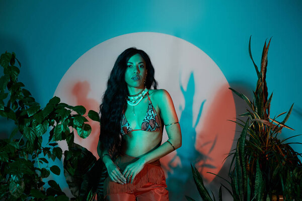 A confident young woman poses in stylish summer attire and jewelry by the plants.