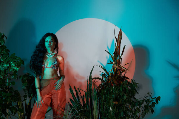 A beautiful young woman poses elegantly against a colorful backdrop with tropical plants around her.