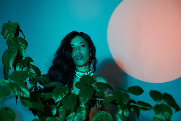 Charming woman with flowing hair poses amid lush plants and colorful lighting during summer.