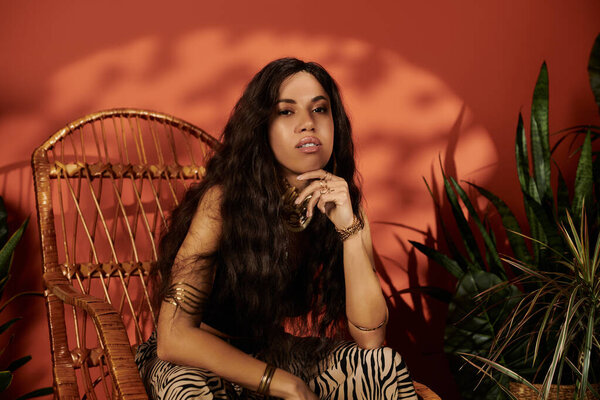 A confident young woman relaxes in a cozy chair, stylishly dressed amidst lush greenery.