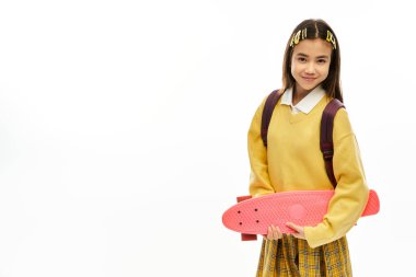 Neşeli bir liseli kız, canlı bir üniformayla, elinde moda bir kaykayla güven içinde poz veriyor..