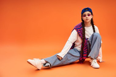 Genç kız bir stüdyoda moda anlayışını sergiliyor, turuncu arka planda gelişigüzel oturuyor..