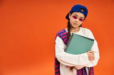Modaya uygun genç bir kız, elinde bir defter, elinde şapka ve güneş gözlüğü ile özgüvenle ayakta duruyor..