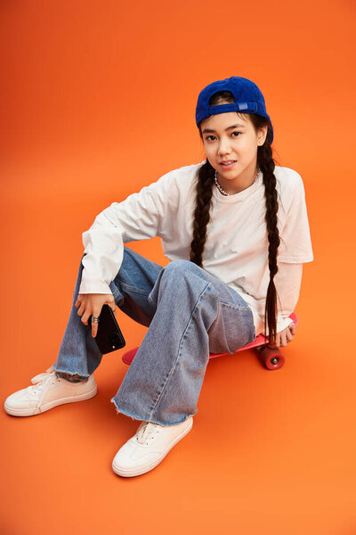 Стильная девушка-подросток сидит на скейтборде, демонстрируя свой модный наряд в студии.