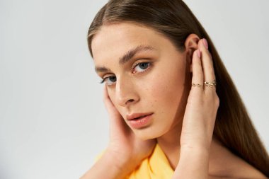 Genç bir kadın, minimalist bir geçmişe karşı poz verirken saçlarına dokunarak güven yayıyor..