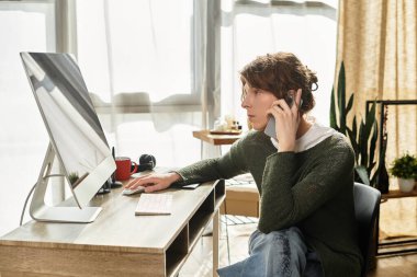 Genç adam masa başında çalışıyor, bilgisayar kullanıyor ve rahat bir evde telefonla konuşuyor..