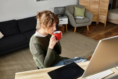 Genç bir adam masada oturmuş, kırmızı bardaktan yudumluyor, çalışırken bilgisayarına odaklanmış..