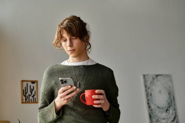 Genç bir öğrenci evinde, elinde kırmızı bir kupayla akıllı telefonunu dikkatle kontrol ederken dinleniyor..