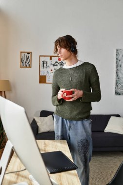 Odaklanmış genç bir öğrenci rahat bir odada durur, bir yudum içer ve teknolojiyi çalışmak için kullanır..