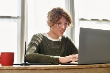 Genç bir adam rahat bir masada otururken, laptopuyla etkileşime girerek derslerine konsantre oluyor..