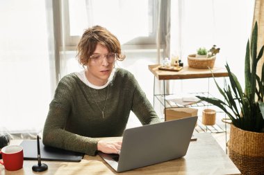 Odaklanmış genç bir adam bilgisayarında çalışıyor. Parlak, bitki dolu bir odada, ahşap bir masada..