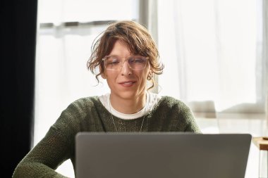 Genç bir adam evde rahat bir şekilde oturur, etkin bir şekilde çalışırken laptopuna odaklanır..