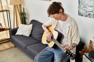 Odaklanmış genç bir adam, sakin bir atmosferde akustik gitar tıngırdatarak bir koltuğa oturur..