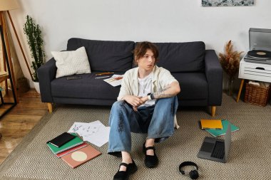 Genç bir öğrenci çalışma materyalleri ve etrafa yayılmış bir dizüstü bilgisayarla rahatça yerde oturuyor..