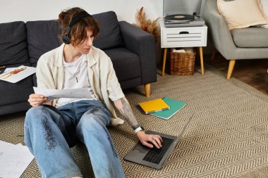 Kendini işine adamış genç bir adam, notlar ve rahat bir ortamda dizüstü bilgisayarla yerde ders çalışıyor..