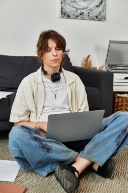 Genç bir adam yerde rahat bir şekilde oturur. Dizüstü bilgisayar kullanırken derslerine dalmıştır..