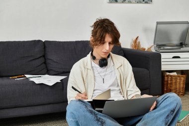 Genç adam evde rahat bir ortamda otururken bir dizüstü bilgisayar ve notlarla meşgul oluyor..