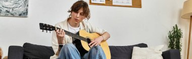Genç bir adam evdeki bir kanepede rahat bir şekilde oturur, gitar çalar ve müziğe odaklanır..