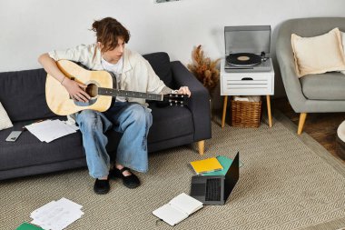 Genç bir öğrenci kanepede gitar çalar. Etrafı kağıtlarla ve rahat bir evde dizüstü bilgisayarla çevrilidir..