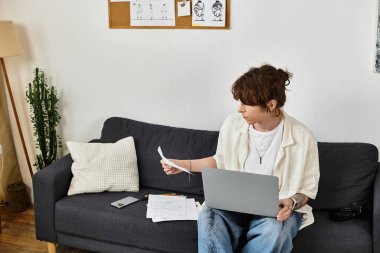 Odaklanmış genç adam, çalışmalarını evde yürütüyor. Kağıtları dengeliyor ve rahat bir ortamda dizüstü bilgisayar kullanıyor..