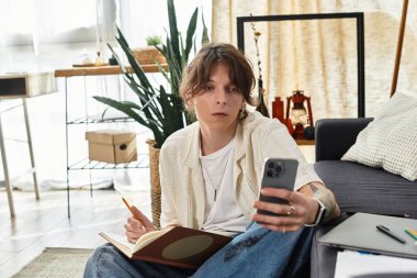 Genç adam rahat bir ev atmosferiyle çevrili telefonunda çalışma materyallerini araştırıyor..