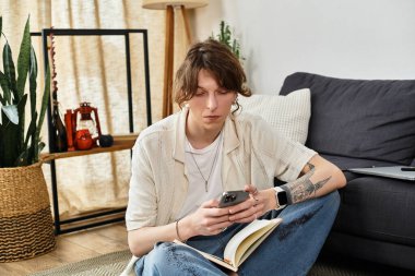 Genç adam evinde derin düşünceler içinde bir kitap ve telefonla rahatça yerde oturuyor..