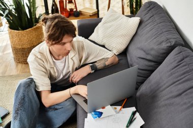 Odaklanmış genç adam evdeki bilgisayarında çalışıyor. Etrafı bitkilerle çevrili ve çalışma materyalleriyle dolu..