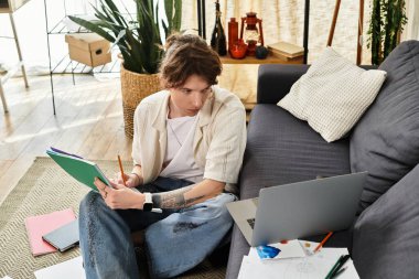 Evde ders çalışan genç bir adam. Etrafı çalışma materyalleriyle çevriliyken laptoptan notlar alıyor..