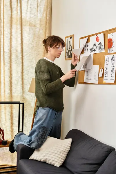 Genç bir adam, tablosunu sıcak bir atmosferle çevrili bir ilan tahtasına iğneleyen bir kanepede duruyor..