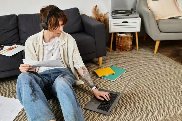 Kendini işine adamış genç bir adam, notlar ve rahat bir ortamda dizüstü bilgisayarla yerde ders çalışıyor..