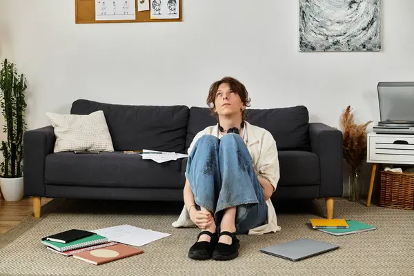 Genç bir öğrenci evde yerde oturup dağınık çalışma materyallerinin arasında düşünüyor..